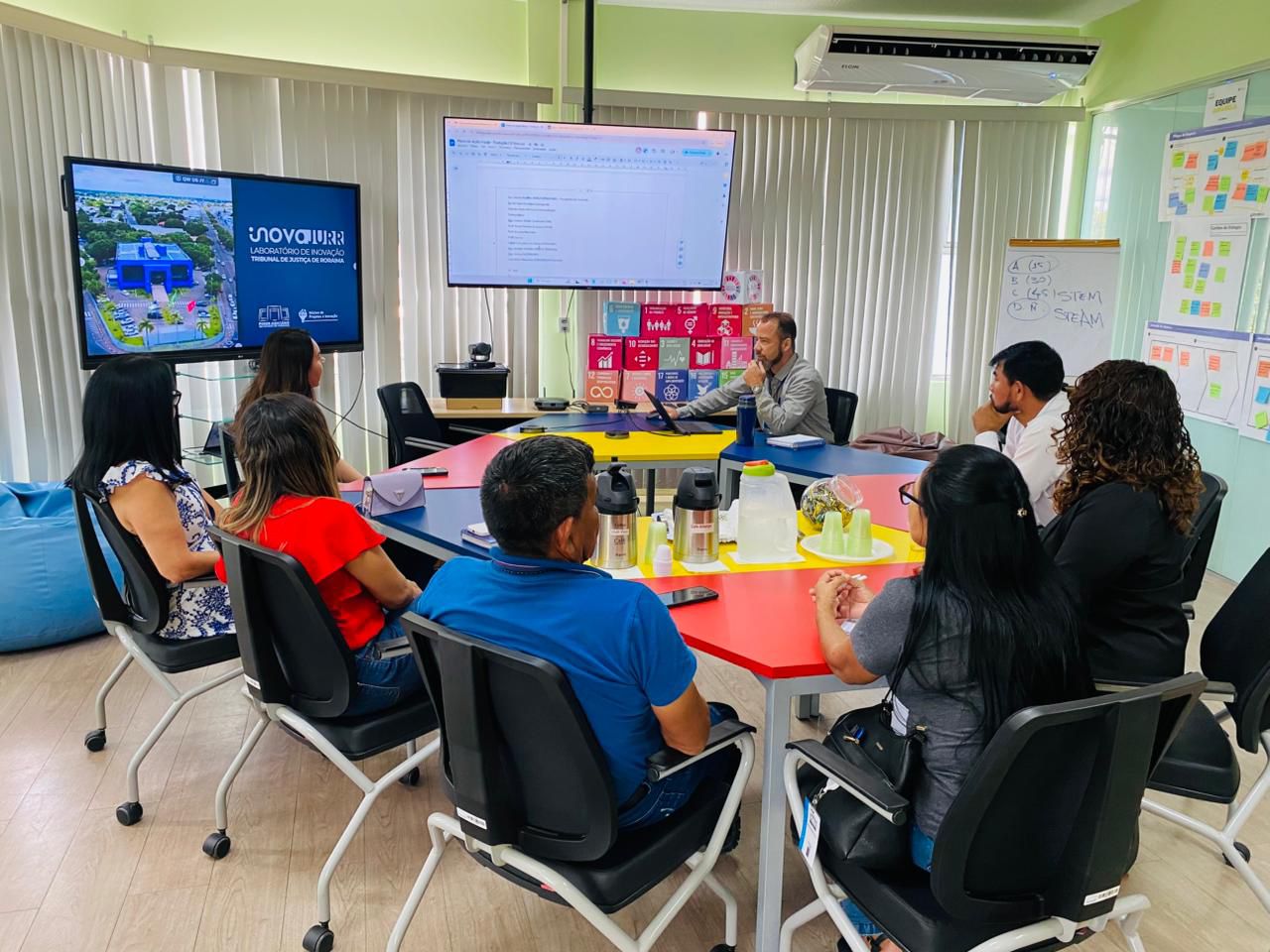 Uma reunião  está acontecendo no laboratório de inovação ou sala de treinamento. Um grupo de cerca de oito pessoas está sentado em torno de uma mesa hexagonal colorida, olhando para um homem no centro que está interagindo com uma grande tela. As telas exibem um slide com o logotipo "INOVARJURR" (Laboratório de Inovação do Tribunal de Justiça de Roraima) e o outro parece ter um documento de texto ou planilha. Há cartazes com os Objetivos de Desenvolvimento Sustentável (ODS) da ONU na parede de trás, sugerindo um foco em inovação e planejamento.