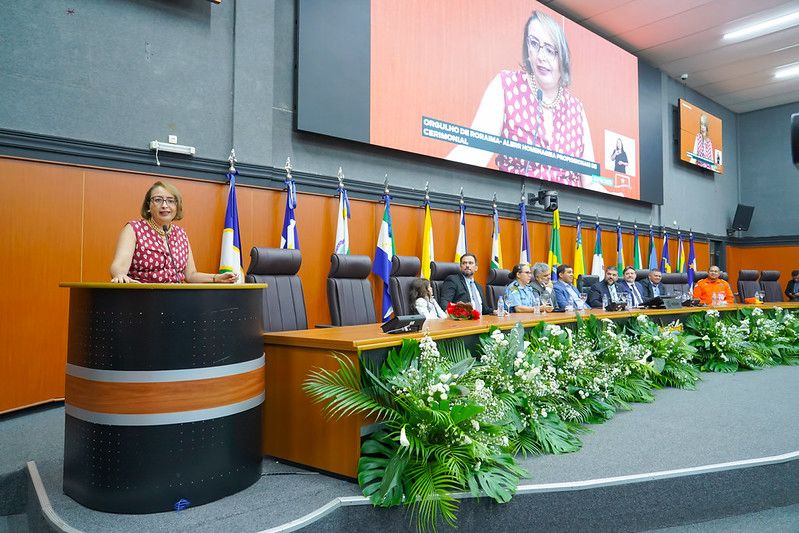  Foto 2: Imagem colorida da Solenidade de entrega da Comenda "Orgulho de Roraima" com a Juíza Auxiliar da presidência Lana Leitão, posando para fotografia falando no púlpito. A mesa diretora conta com diversas autoridades.