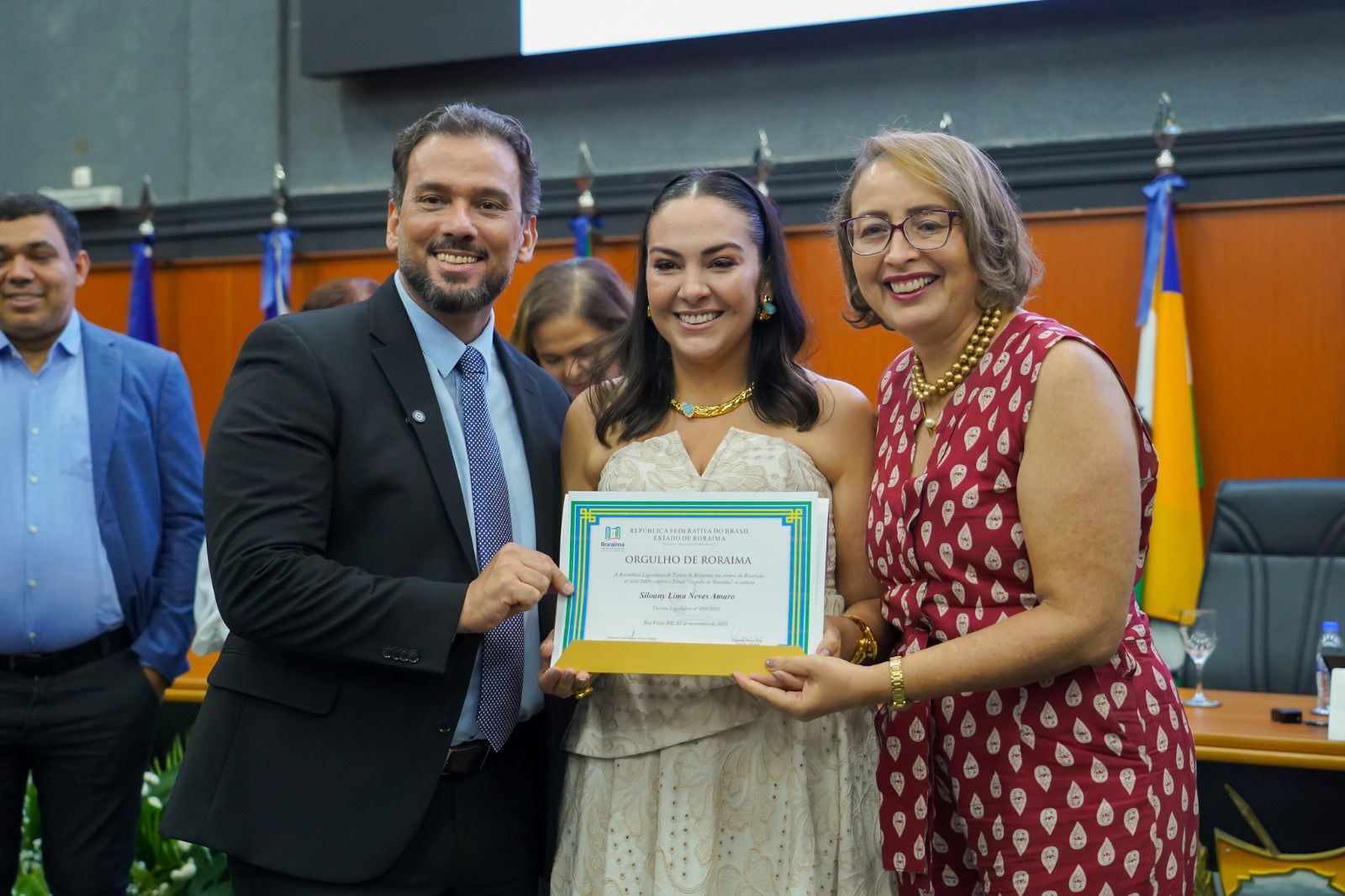 Foto 1: Imagem colorida de três pessoas posando para fotografia com o certificado em mãos durante a solenidade. 