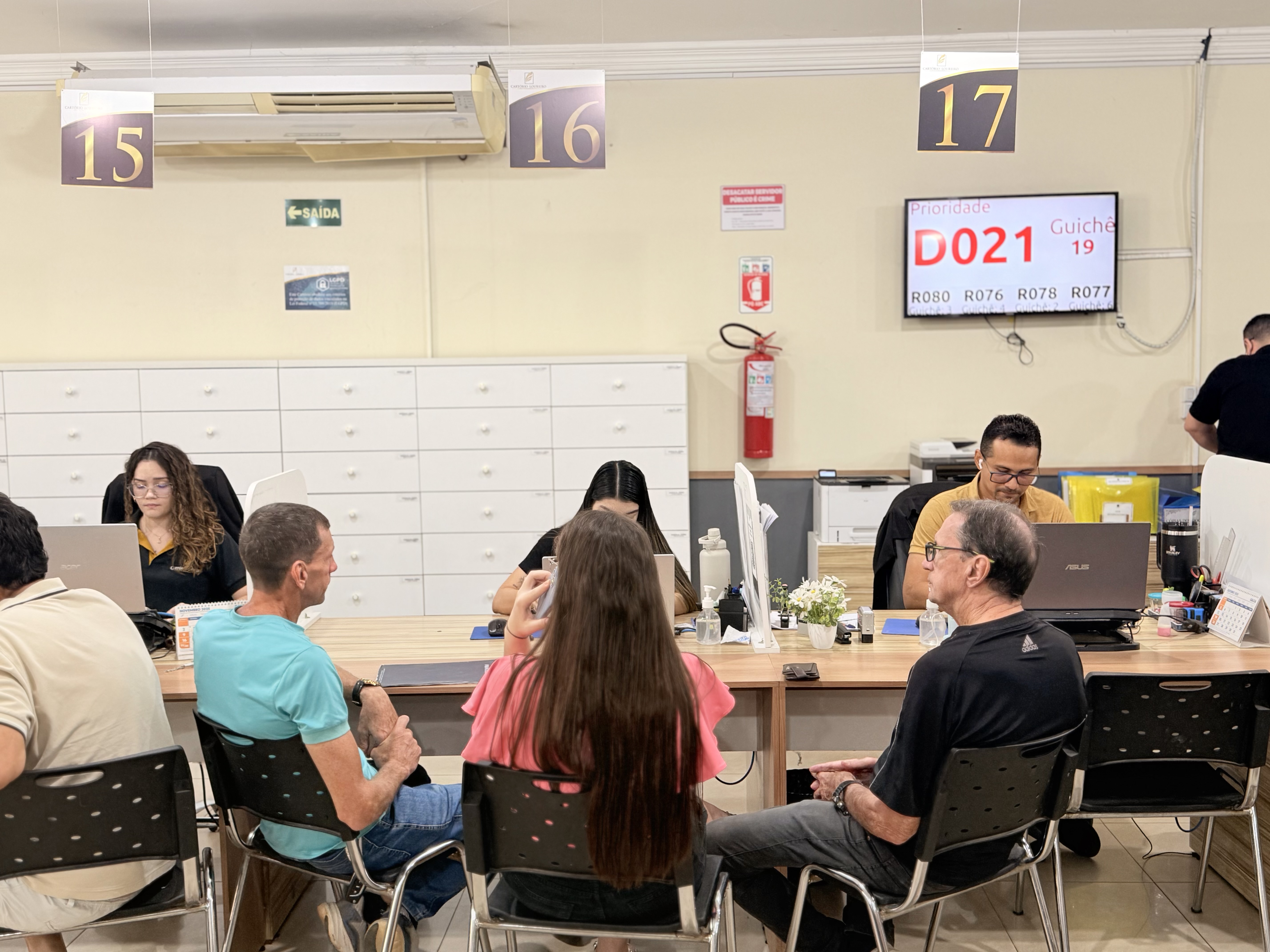 Imagem colorida mostra um ambiente de atendimento ao público. Quatro guichês estão sendo ocupados por atendentes (três mulheres e um homem). Cinco pessoas (três homens e duas mulheres) estão sentadas em frente ao balcão sendo atendidas. No fundo, números grandes indicam os guichês (15, 16, 17) e um monitor mostra a senha atual "D021" para o guichê "19". Imagem colorida mostra um ambiente de atendimento ao público. Quatro guichês estão sendo ocupados por atendentes (três mulheres e um homem). Cinco pessoas (três homens e duas mulheres) estão sentadas em frente ao balcão sendo atendidas. No fundo, números grandes indicam os guichês (15, 16, 17) e um monitor mostra a senha atual "D021" para o guichê "19".