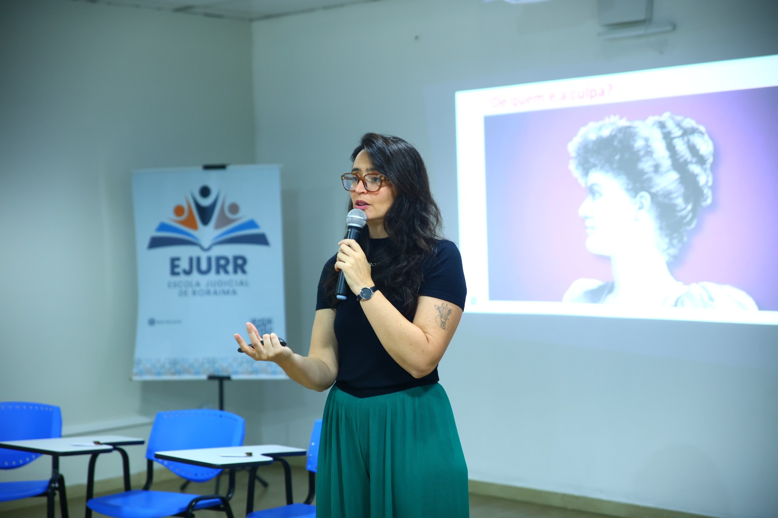  imagem dando close na palestrante em um momento de fala, segurando um microfone ao fundo há o banner da ejurr.