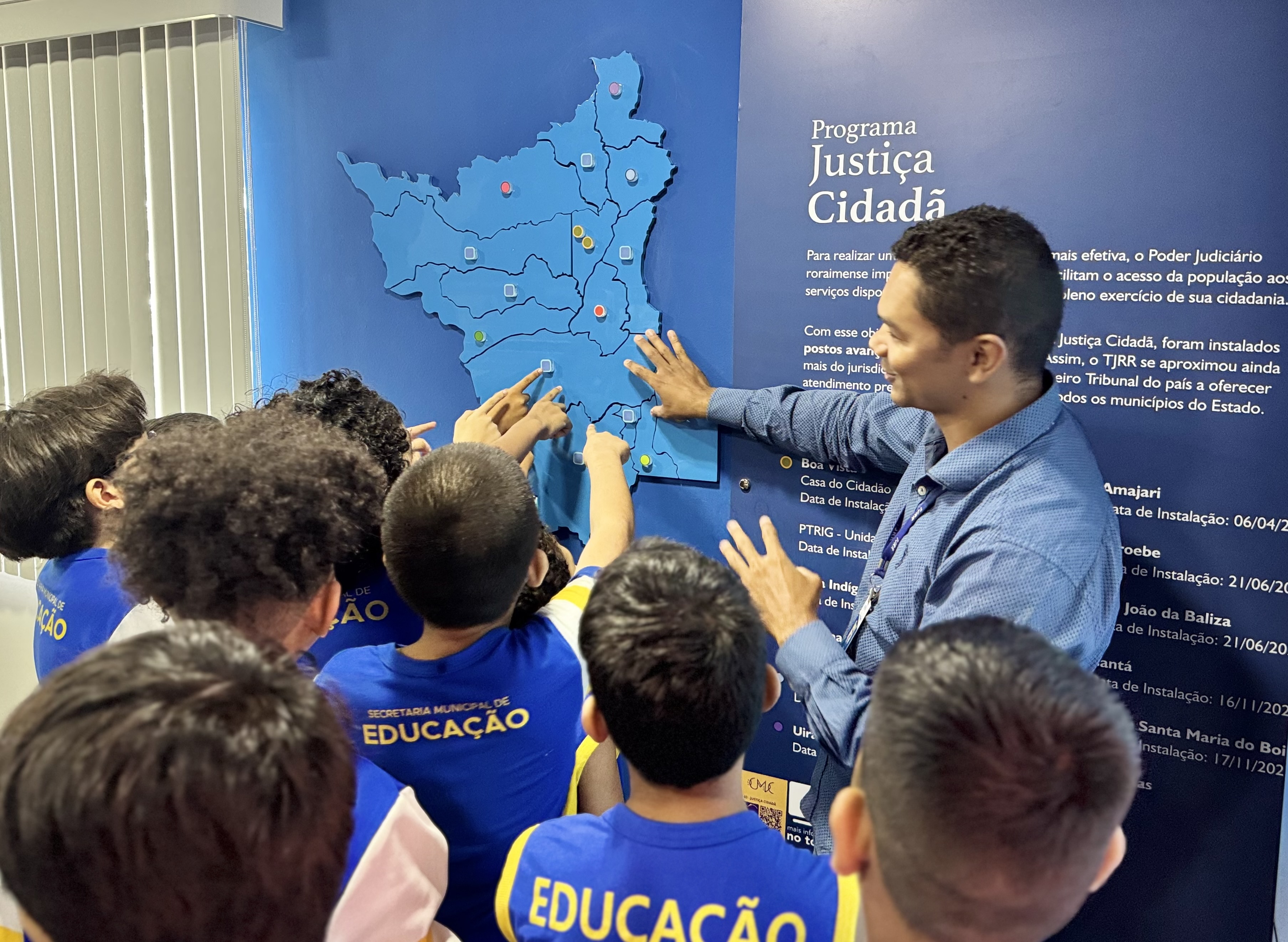 Imagem colorida mostra um grupo de crianças de camiseta azul e amarelo, acompanhadas por um homem adulto. Elas estão reunidas em frente a um painel azul com a ilustração de um mapa do estado de Roraima, apontando para diferentes regiões. Imagem colorida mostra um grupo de crianças de camiseta azul e amarelo, acompanhadas por um homem adulto. Elas estão reunidas em frente a um painel azul com a ilustração de um mapa do estado de Roraima, apontando para diferentes regiões.