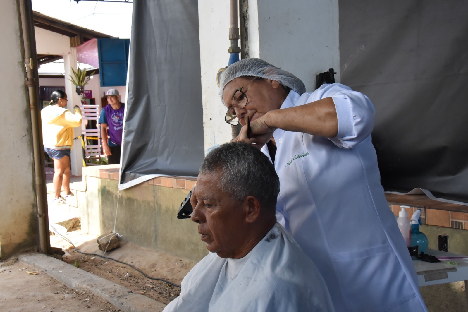 Um cabeleireiro corta o cabelo de um homem em um ambiente externo, enquanto outras pessoas estão ao fundo. Um cabeleireiro corta o cabelo de um homem em um ambiente externo, enquanto outras pessoas estão ao fundo.