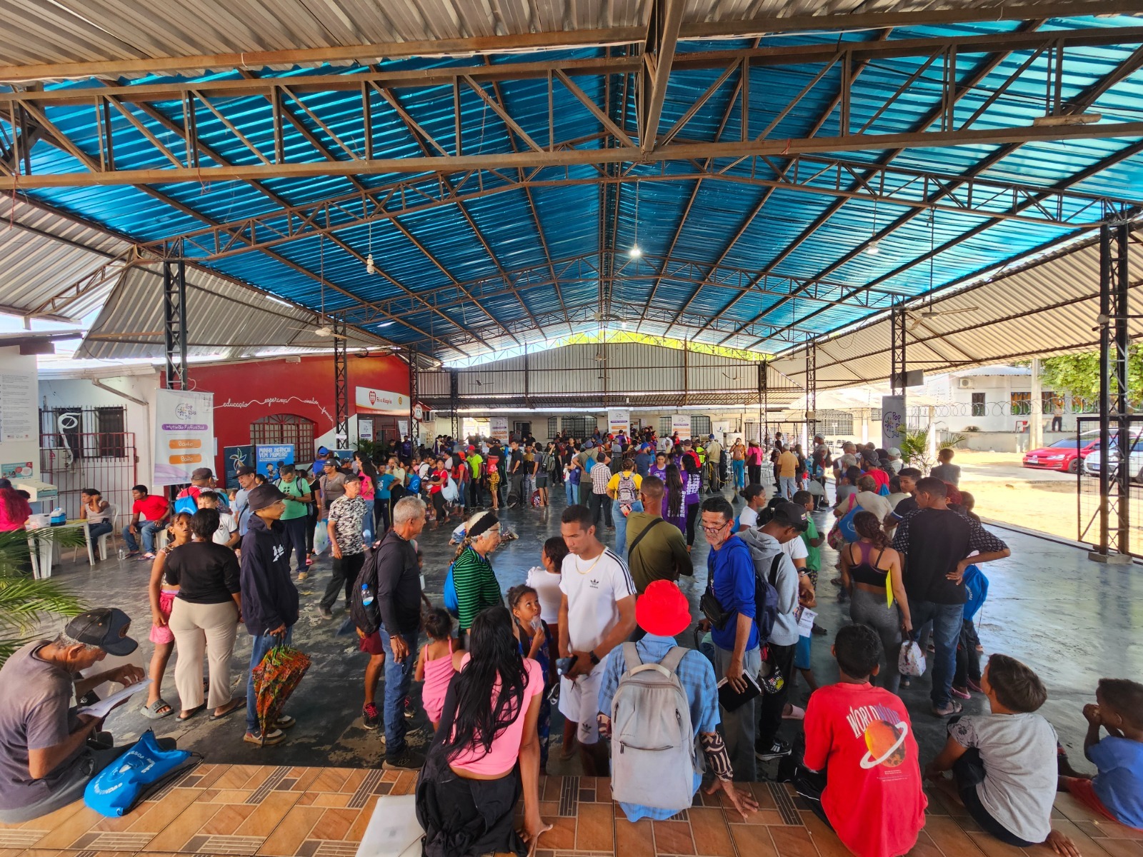 Multidão de pessoas em um espaço coberto, aguardando em filas. O ambiente apresenta um teto azul e paredes com informações visíveis ao fundo. Multidão de pessoas em um espaço coberto, aguardando em filas. O ambiente apresenta um teto azul e paredes com informações visíveis ao fundo.