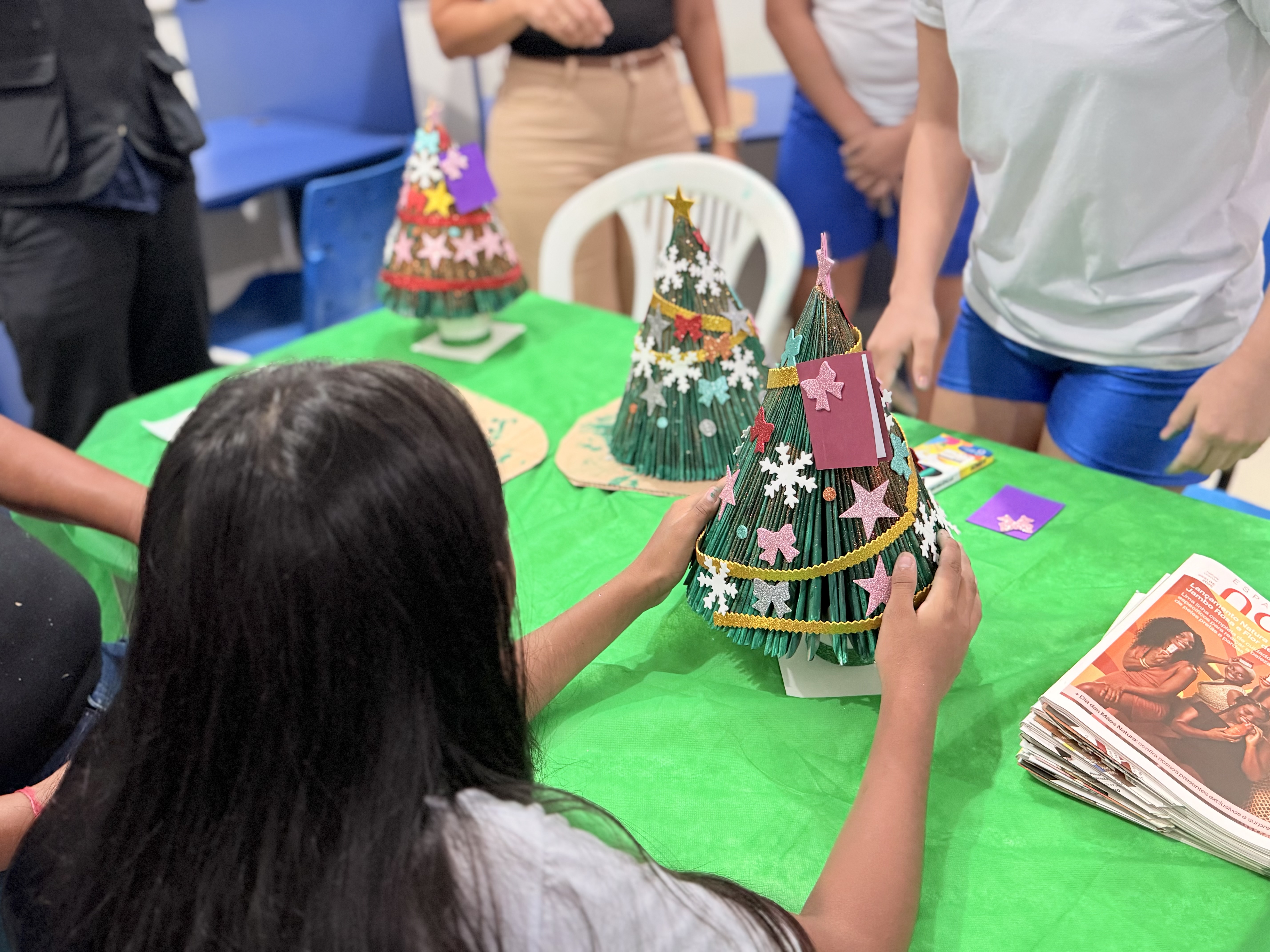 Imagem colorida mostra uma pessoa sentada à mesa verde, segurando uma árvore de Natal artesanal decorada com estrelas, laços e enfeites coloridos, feita de papel ou material reciclado. Outras pessoas e árvores semelhantes estão ao redor, sobre a mesa. Há um monte de revistas no canto direito.