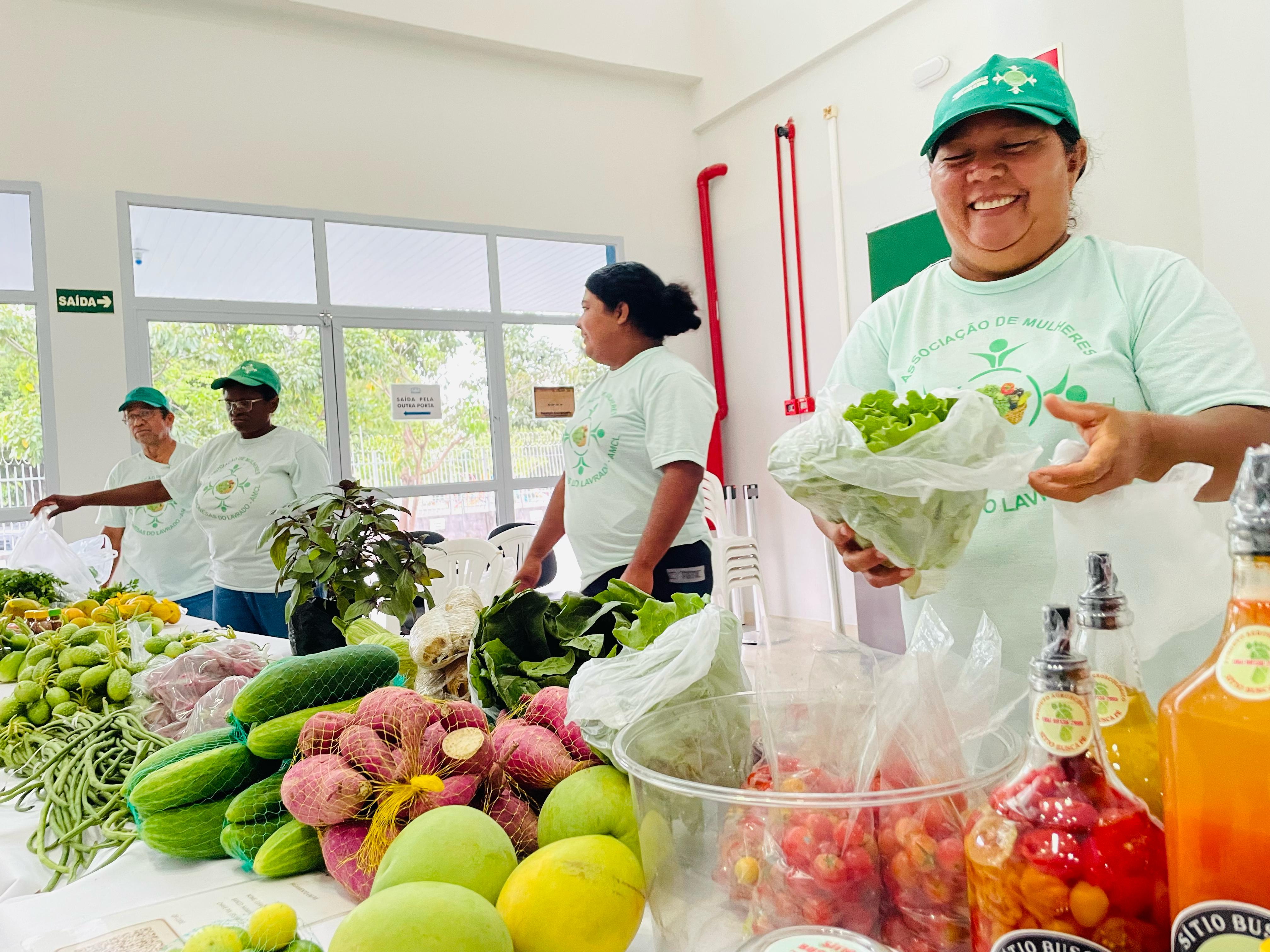Imagem colorida mostra uma mesa repleta de hortaliças, frutas, legumes e frascos de produtos artesanais, como conservas e sucos. Três mulheres vestem camiseta branca e boné verde com o logo da "Associação de Mulheres do Lavrado", estando atrás da mesa, uma delas segurando um pacote de alface. Ao fundo, há cadeiras brancas e uma parede com janelas de vidro, além de placas de saída. O ambiente é bem iluminado e aparenta ser interno.