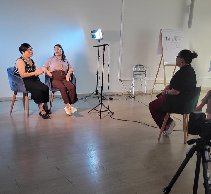 imagem colorida de um momento de gravação onde há duas mulheres sentadas uma ao lado da outra e a frente uma mulher entrevistando também sentada. 