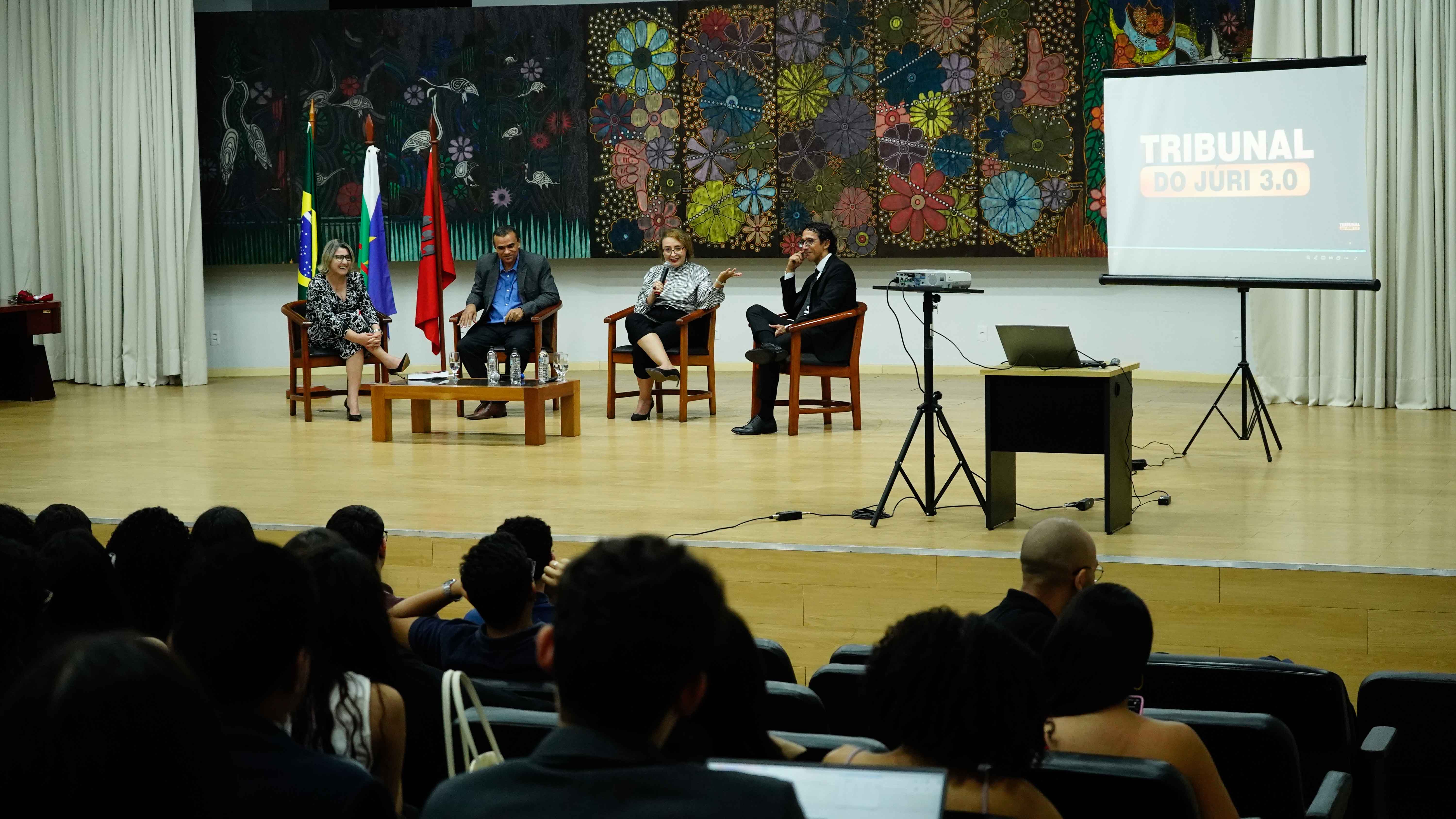 Auditório com plateia assistindo a quatro pessoas sentadas em cadeiras no palco, participando de uma mesa-redonda. Ao fundo, há um grande painel colorido com flores e bandeiras posicionadas à esquerda. À direita, um telão projeta o título “Tribunal do Júri 3.0”.