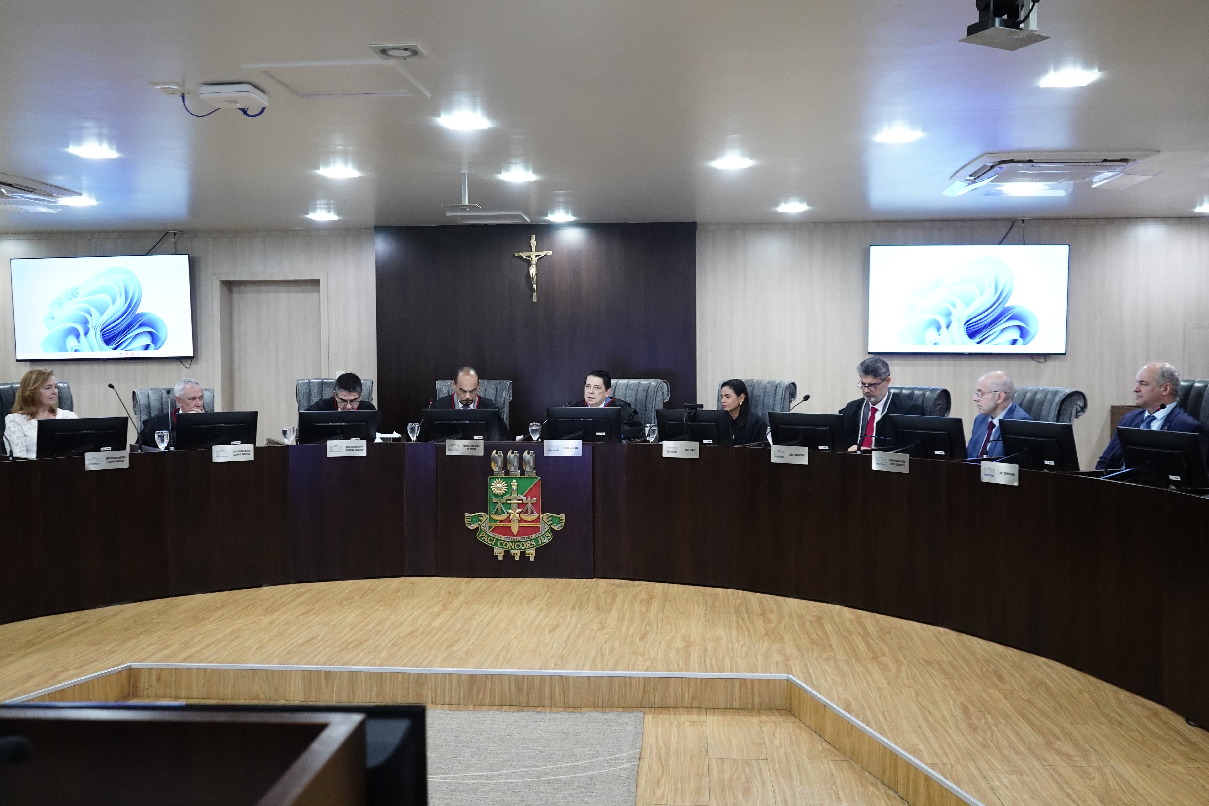  Imagem 2: Imagem colorida mostra nove autoridades do Judiciário roraimense sentados no plenário do Tribunal Pleno do TJRR. Há duas tv 's atrás do plenário.