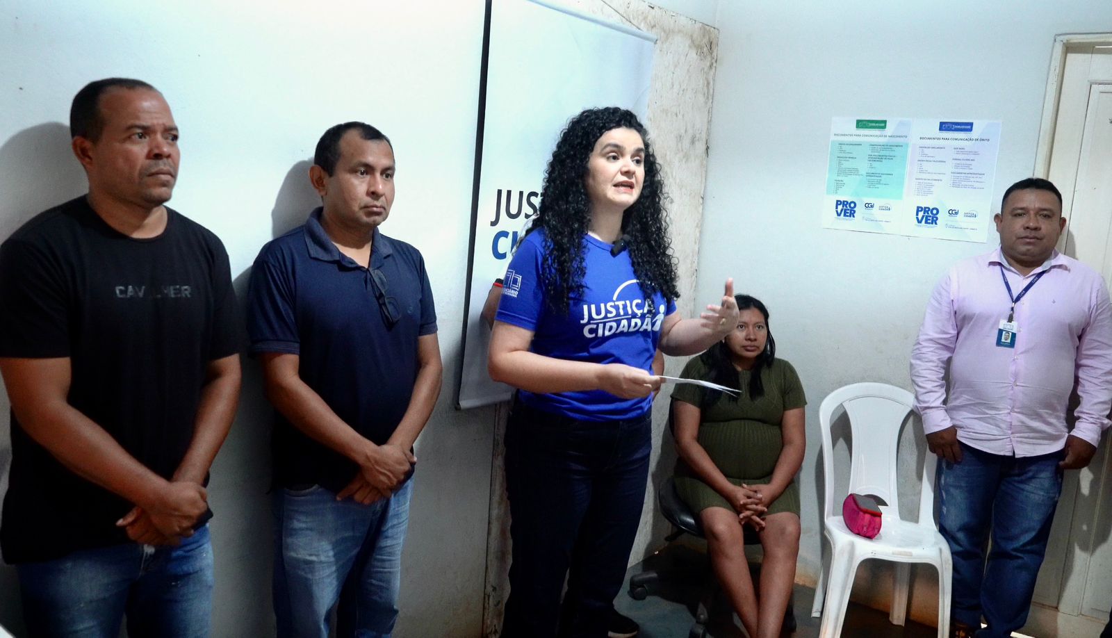 imagem colorida mostra um grupo de cinco pessoas, três homens e duas mulheres, em pé e sentados em uma sala. Uma mulher no centro, vestindo uma camiseta azul com a estampa "Justiça Cidadã" e cabelo cacheado, está falando e gesticulando com a mão direita, enquanto segura um papel na esquerda. Dois homens estão em pé à esquerda dela e um homem está em pé à direita. Uma mulher está sentada em uma cadeira plástica branca à direita da oradora. Um banner azul e um cartaz informativo estão visíveis ao fundo. imagem colorida mostra um grupo de cinco pessoas, três homens e duas mulheres, em pé e sentados em uma sala. Uma mulher no centro, vestindo uma camiseta azul com a estampa "Justiça Cidadã" e cabelo cacheado, está falando e gesticulando com a mão direita, enquanto segura um papel na esquerda. Dois homens estão em pé à esquerda dela e um homem está em pé à direita. Uma mulher está sentada em uma cadeira plástica branca à direita da oradora. Um banner azul e um cartaz informativo estão visíveis ao fundo.