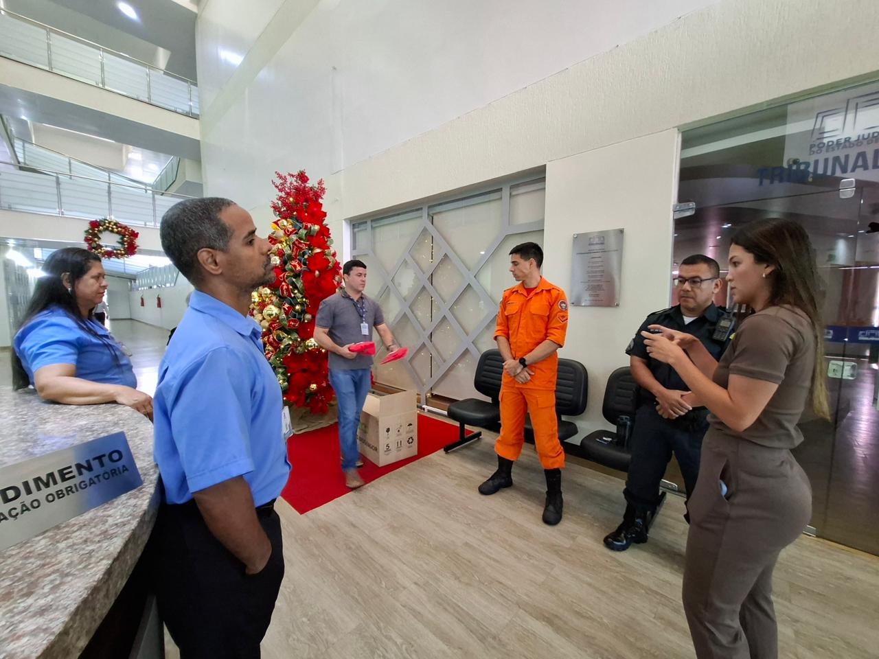 Imagem colorida mostra um ambiente decorado para o Natal, com uma grande árvore vermelha ao fundo. Um homem com crachá segura pacotes vermelhos próximos a uma caixa de papelão no chão, enquanto outras pessoas em diferentes uniformes, incluindo um de roupa laranja e outro de preto, o observam. À esquerda, uma mulher se apoia em um balcão.