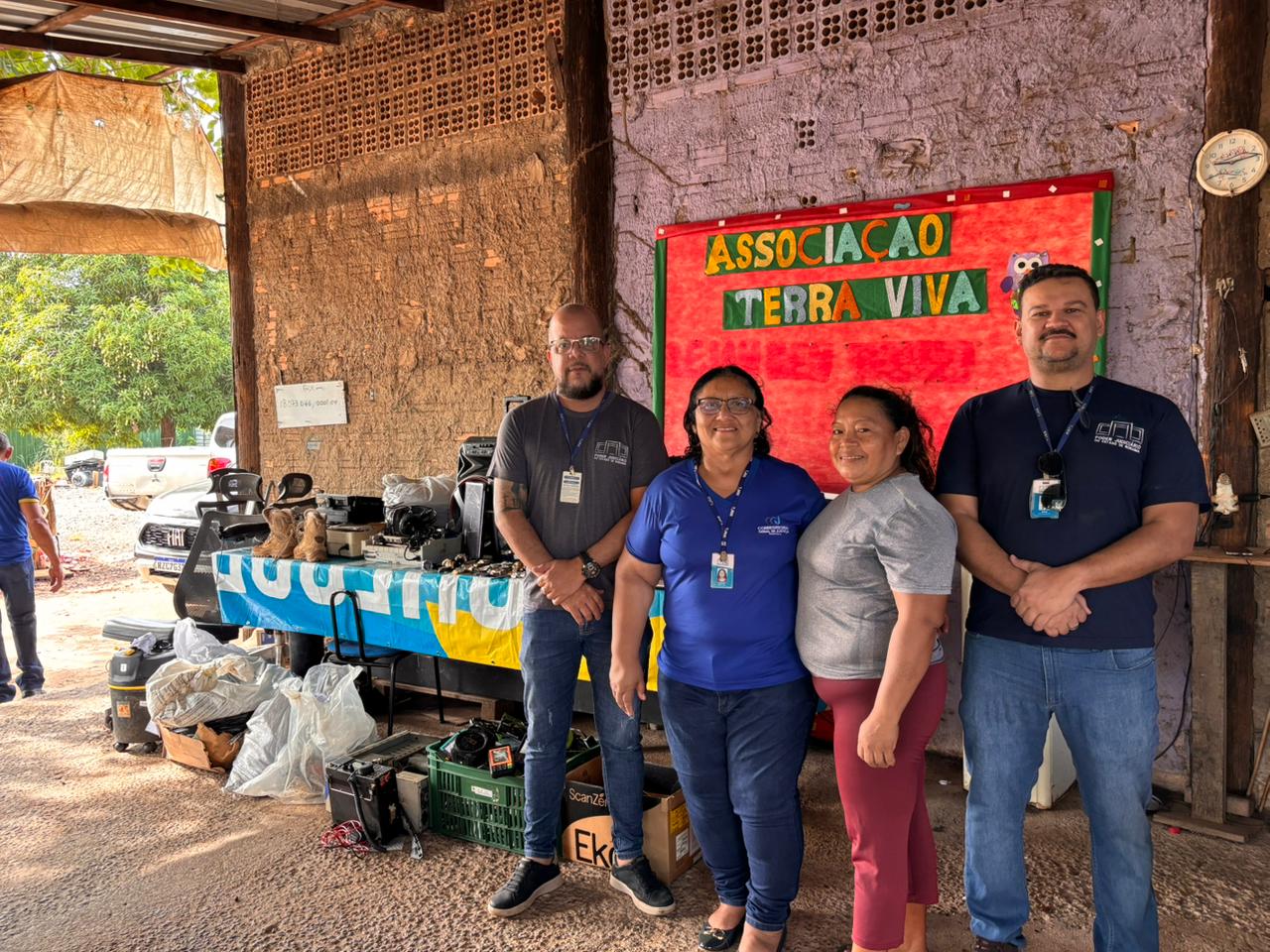 Quatro pessoas posam em pé em frente ao espaço da Associação Terra Viva. Ao fundo, há uma mesa com diversos equipamentos eletrônicos apreendidos. Na parede, um cartaz colorido com o nome da associação identifica o local.