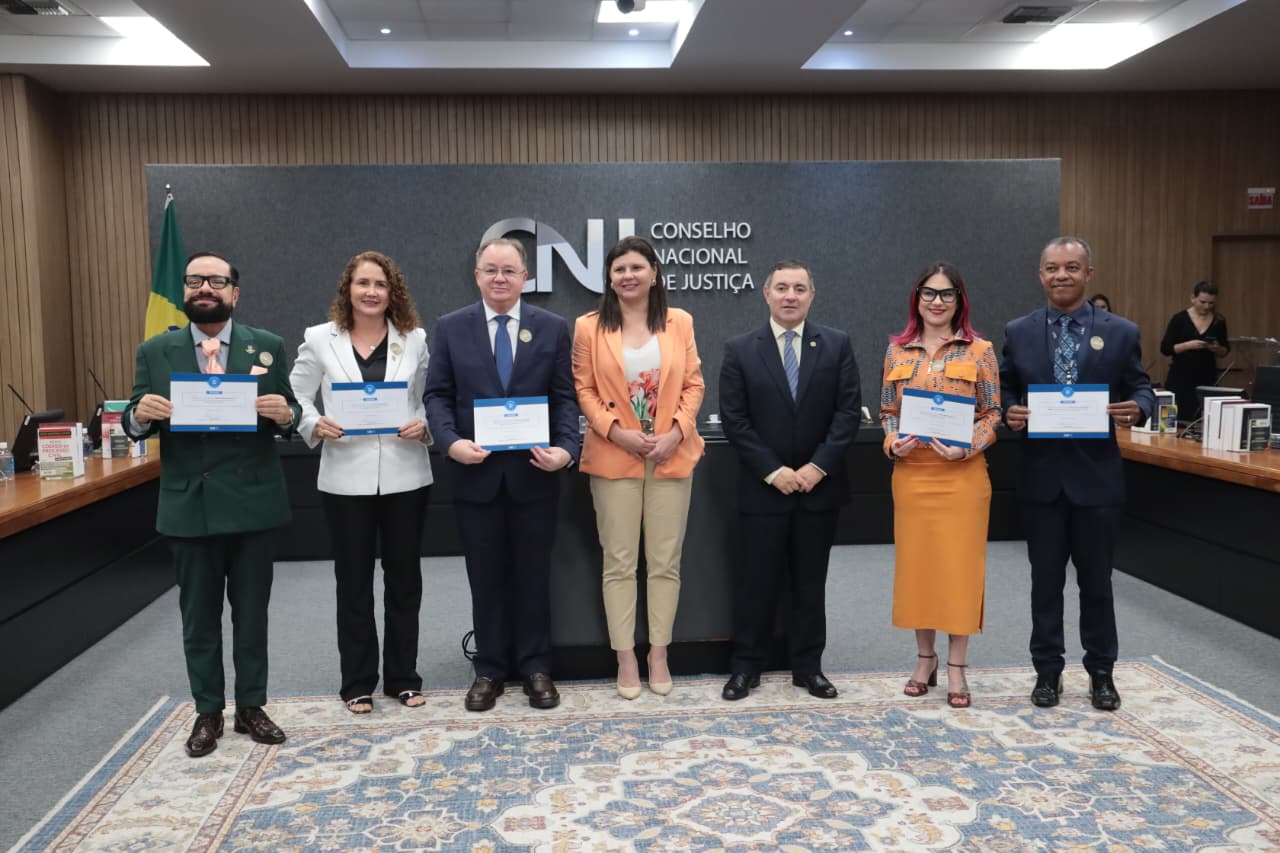 Uma foto colorida mostra uma cerimônia solene, contendo o presidente do TJRR, desembargador Leonardo Cupello, ao lado de seis autoridades do Judiciário brasileiro, que estão alinhadas no centro do auditório, em frente a um grande painel cinza com a logo do CNJ (Conselho Nacional de Justiça). Elas vestem trajes sociais ou uniformes de gala, e a maioria segura certificados ou placas. Uma foto colorida mostra uma cerimônia solene, contendo o presidente do TJRR, desembargador Leonardo Cupello, ao lado de seis autoridades do Judiciário brasileiro, que estão alinhadas no centro do auditório, em frente a um grande painel cinza com a logo do CNJ (Conselho Nacional de Justiça). Elas vestem trajes sociais ou uniformes de gala, e a maioria segura certificados ou placas.