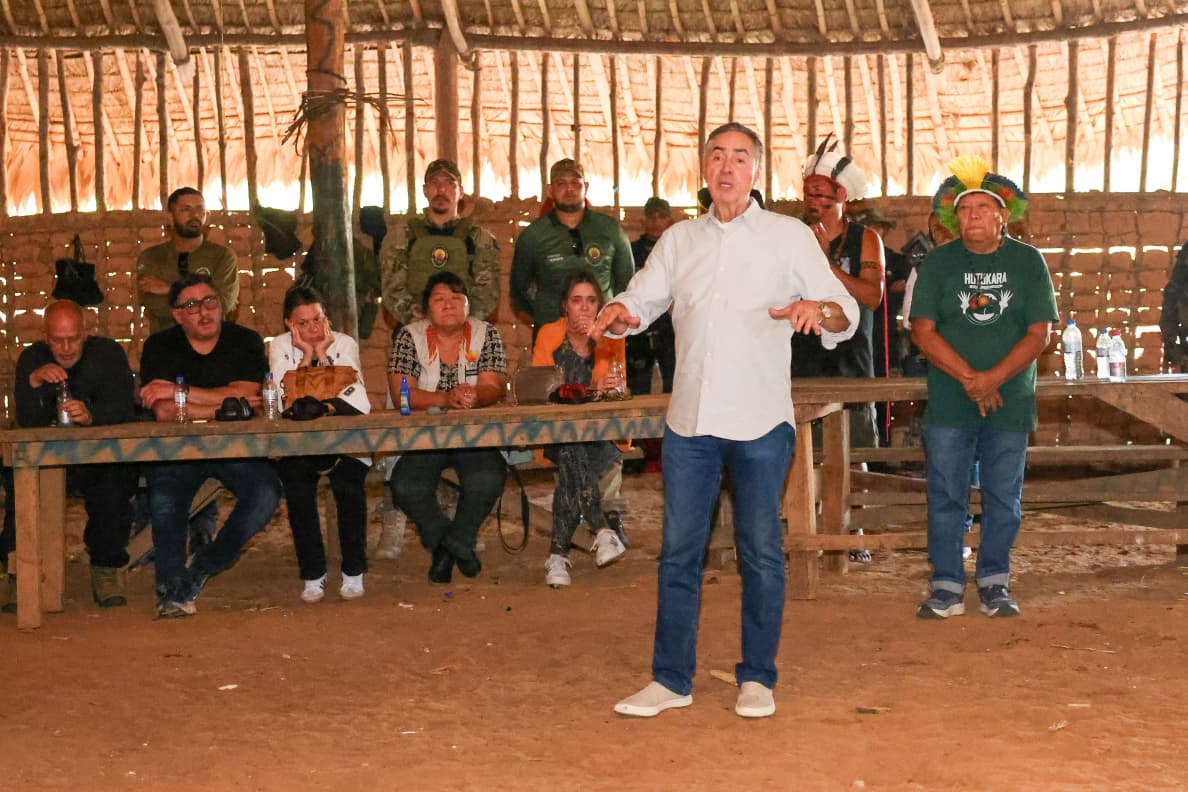 Fotografia mostra um grupo de pessoas dentro de uma grande estrutura de madeira e palha. No primeiro plano, um homem com camisa social branca e calça jeans, está de pé no centro da cena. Ele gesticula enquanto fala. Atrás dele, ao longo de uma mesa, outros indivíduos, incluindo indígenas com cocares e roupas tradicionais, estão sentados. 