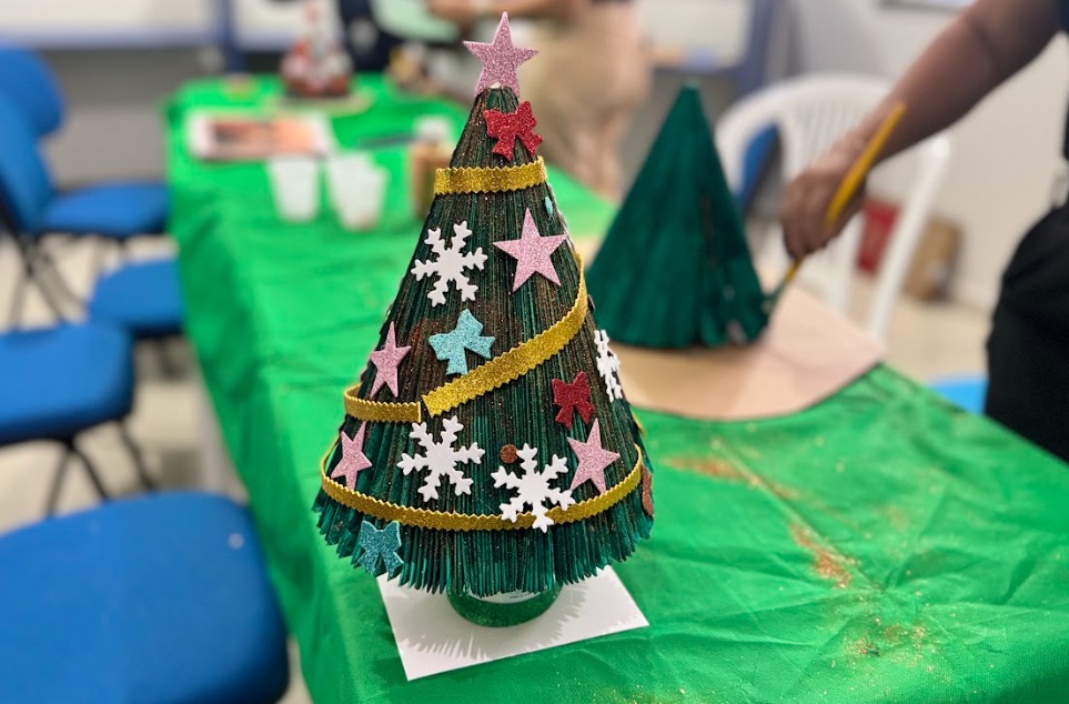 imagem colorida mostra uma mesa coberta com uma toalha verde, onde está exposto um pequeno pinheiro de Natal artesanal em formato de cone. A árvore é decorada com tiras onduladas douradas e diversos enfeites feitos de EVA, como estrelas e flocos de neve nas cores branco, rosa, vermelho, azul e verde. No topo há uma estrela rosa. A base do cone está apoiada sobre um quadrado de papel branco. Ao fundo, desfocado, aparecem cadeiras azuis, copos descartáveis sobre a mesa e outra árvore em construção. À direita, vê-se parcialmente o braço de uma pessoa segurando um pincel amarelo, trabalhando em uma estrutura semelhante. A cena transmite clima de oficina artesanal de Natal.