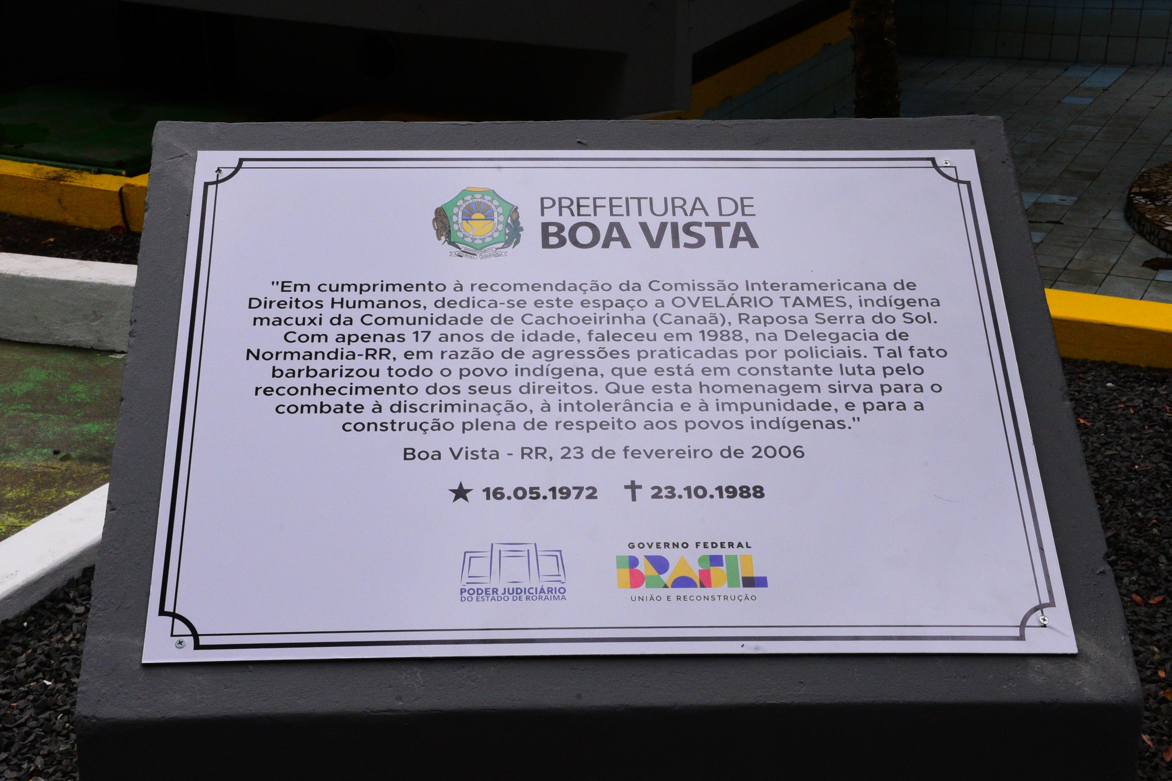 Imagem colorida mostra a placa revitalizada do memorial do indígena Ovelário Tames, na praça do coreto. Abaixo a frase “Homenagem aos Povos Originários - Memorial Indígena revitalizado no Dia dos Direitos Humanos reforça luta por Justiça em Roraima.”