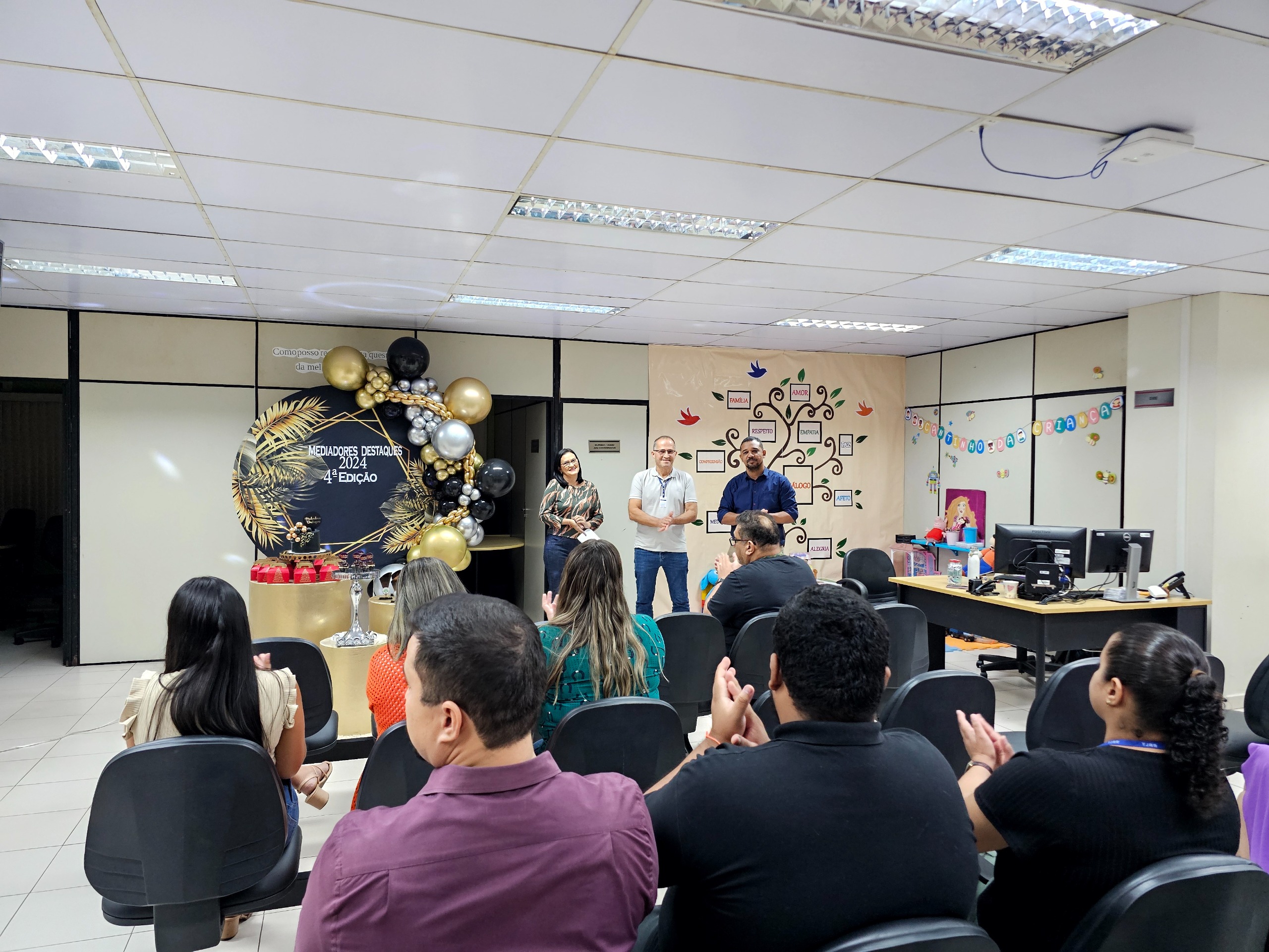 Imagem colorida mostra três servidores em pé apresentando os premiados para os demais servidores que estão sentados em cadeiras assistindo. 