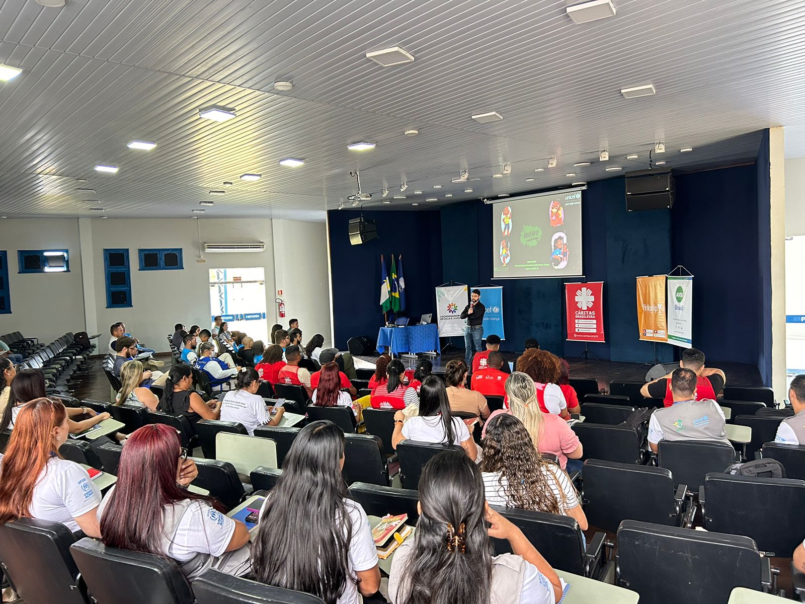 A imagem colorida mostra os participantes da oficina em cadeiras numa sala cheia, todos acompanhando as informações do evento. Abaixo a frase: Oficina reforça Direitos de Crianças e Adolescentes Imigrantes em Roraima