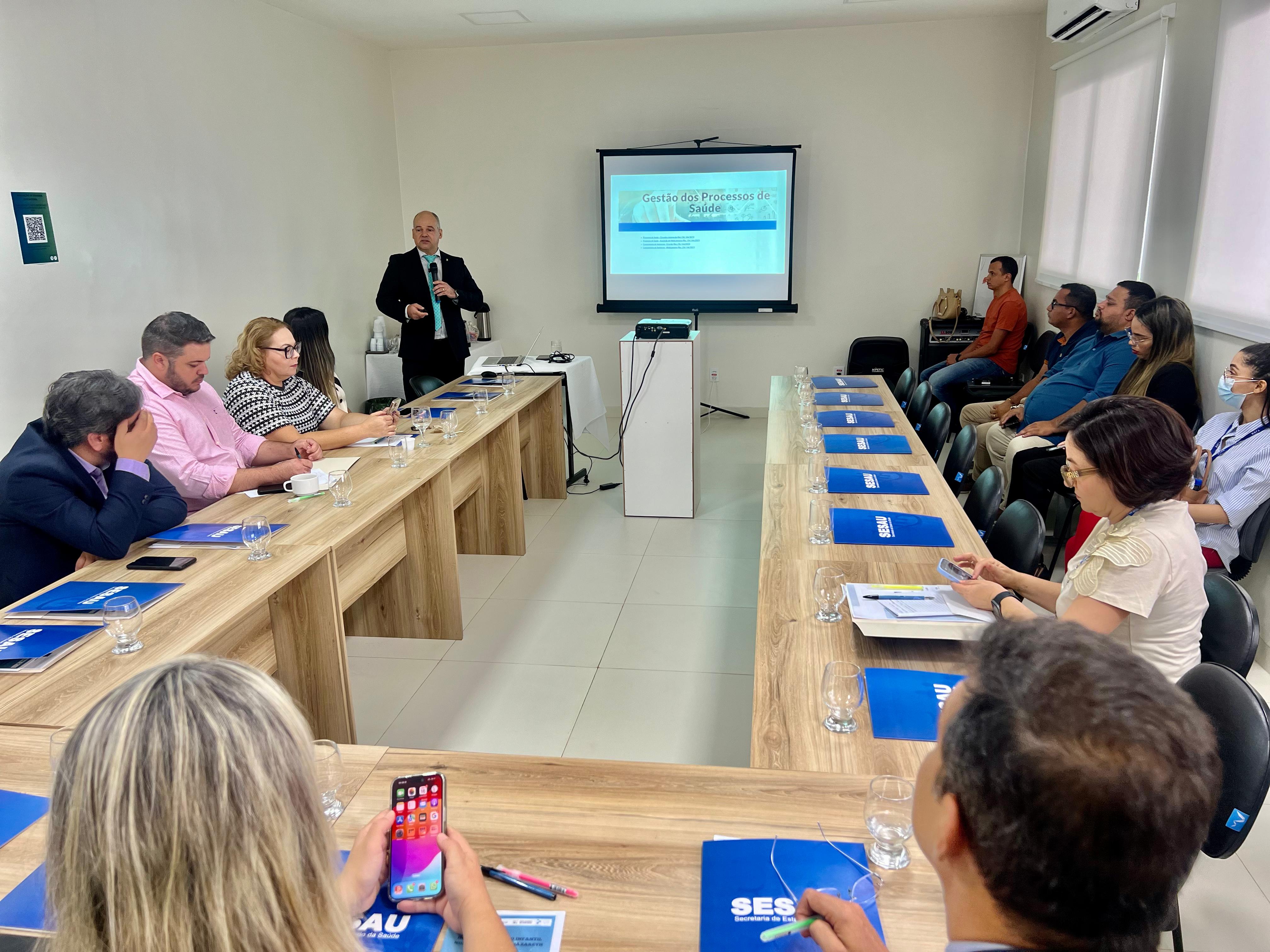 Imagem colorida mostra o juiz Eduardo Carvalho, coordenador do CES/RR, palestrando para público de doze pessoas, entre elas, autoridades da saúde em Roraima, sentados em volta de mesa de reunião posicionada em forma de “U”