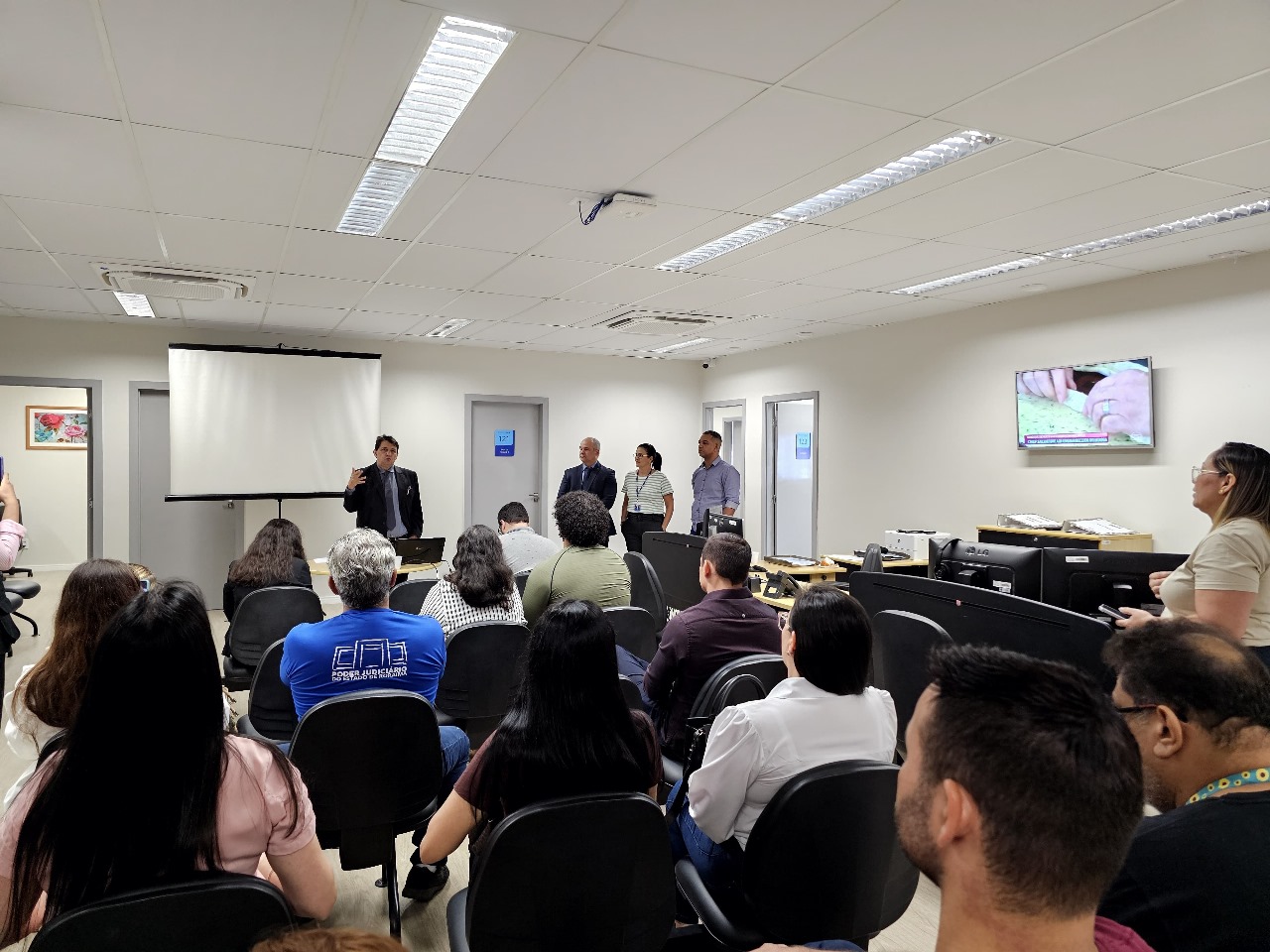  foto colorida mostra uma sala com onze pessoas sentadas e quatro pessoas em pé prestando atenção na fala do Presidente do TJRR, desembargador Jésus Nascimento, ao fundo. Abaixo a frase "APERFEIÇOAMENTO - Nupemec realiza oficina com mediadores e conciliadores judiciais"