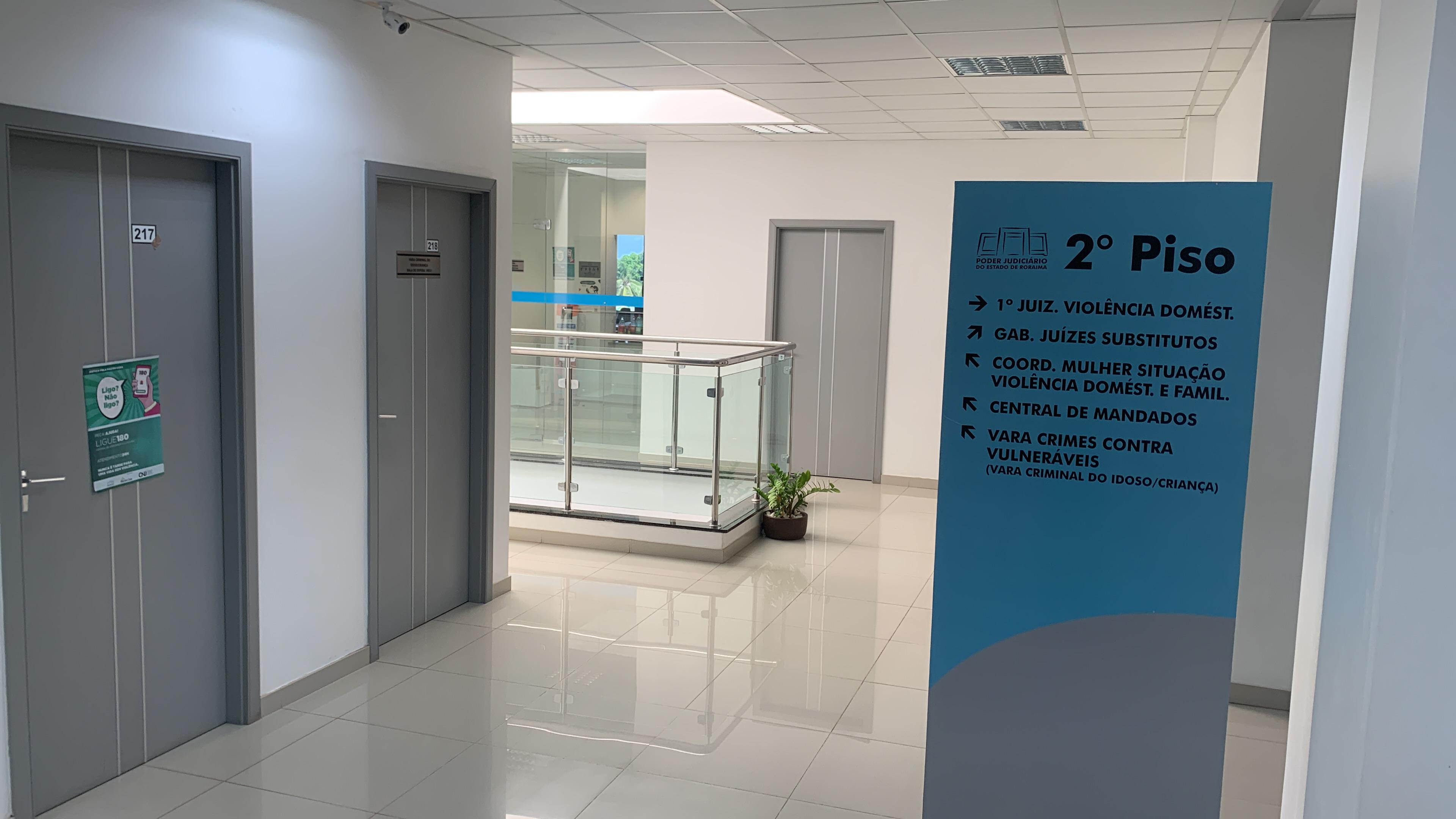 foto colorida do corredor com uma placa de metal e cor azul com  sinalização aos serviços prestados dentre ele está a o 1° Juizado de Violência Doméstica