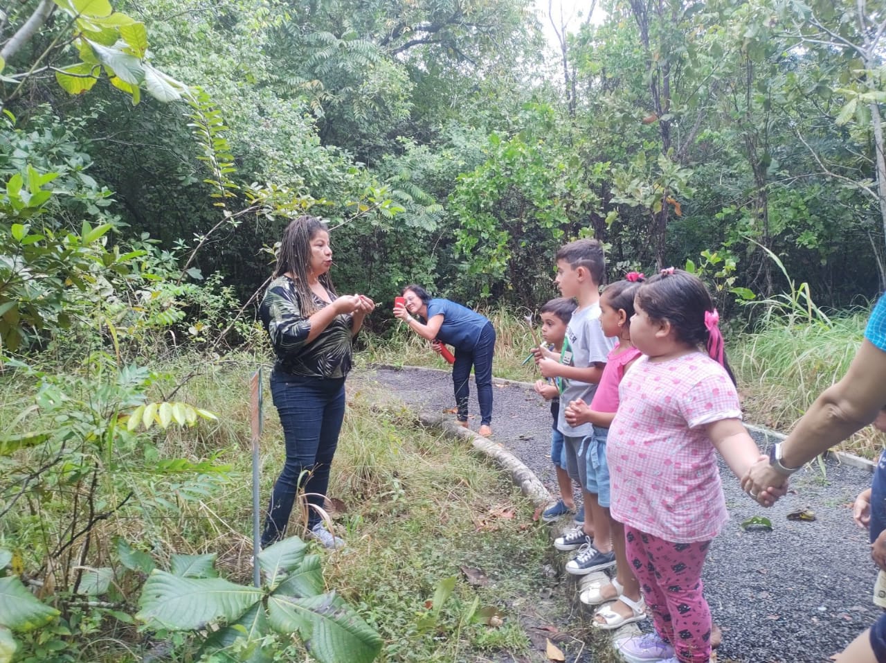  Imagem colorida ilustrativa mostra uma servidora conversando com as crianças, as mesmas estão de mãos dadas no limite da estrada de pedras, ao redor observa-se um fundo com diversos tipos de vegetação.