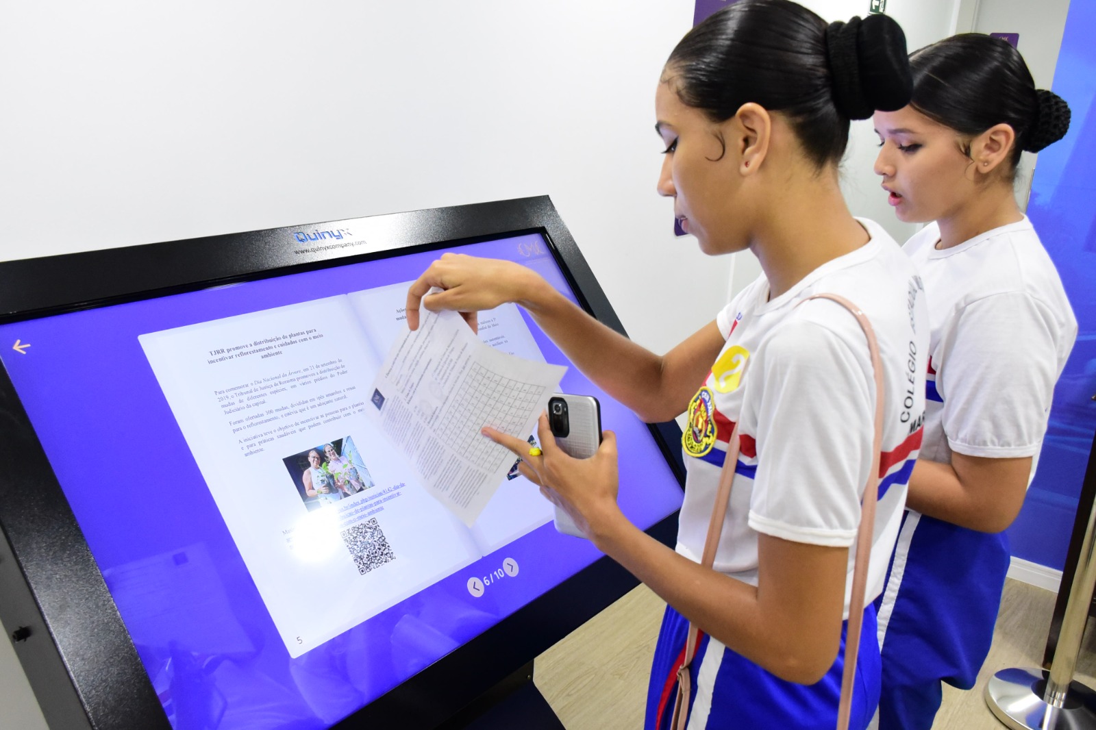 Imagem colorida mostra  duas alunas do Colégio Estadual Militarizado Maria Sônia de Brito Oliva em frente a um painel digital.