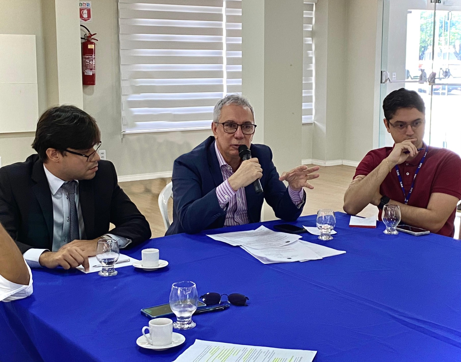 Imagem colorida contém três pessoas sentados em volta de uma mesa, entre elas encontra-se o desembargador e coordenador do GMF/TJRR, Almiro Padilha falando ao microfone. 