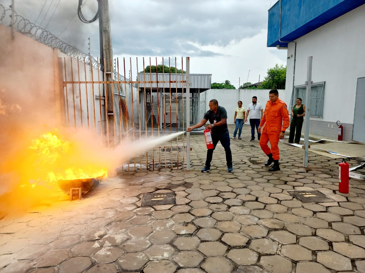 Imagem colorida mostra duas pessoas, sendo um servidor segurando um extintor enquanto apaga o fogo, e um bomeiro posicionado logo atrás do servidor dando apoio técnico durante treimamento do Corpo de Bombeiros. 