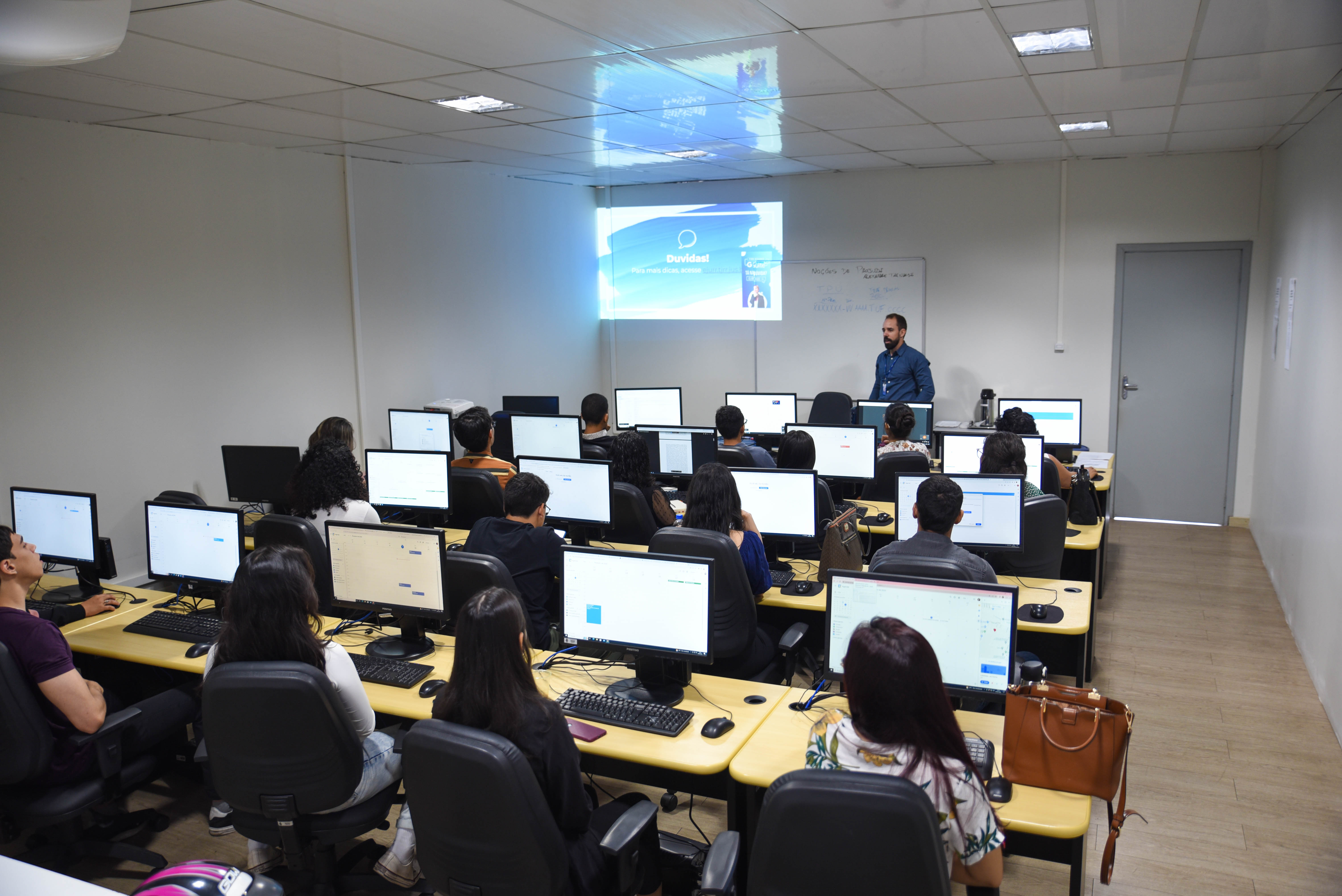  Foto colorida de estudantes sentados em uma sala de aula. Ao fundo, uma professora em pé em frente a um quadro branco e uma projeção na pared