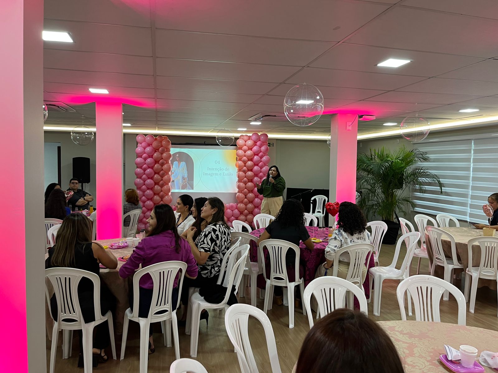 Foto colorida mostra pessoas sentadas às mesas no evento, e ao fundo a palestrante falando