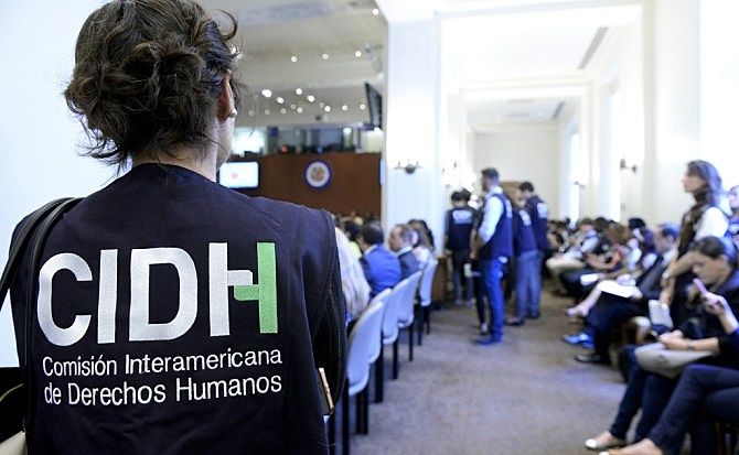 Foto colorida mostrando pessoas sentadas em cadeiras de auditório. Em primeiro plano uma pessoa está usando um colete com a sigla de uma instituição em espanhol: CIDH - Comisión Interamericana de Derechos Humanos