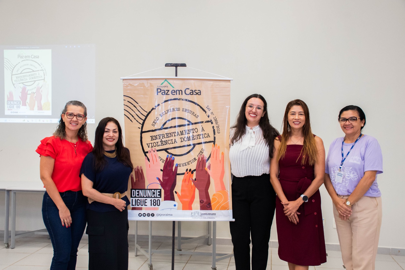 Foto colorida mostra cinco mulheres da CEVID/TJRR posando para a foto ao lado do banner do Selo Mulheres Seguras. Entre elas, a juiza Suelen Alves